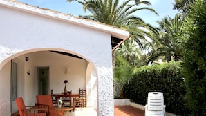 A white building with a tiled patio, wooden furniture, and a white plastic chair. - Villa LA TARDE 1ª Línea Playa (Denia)