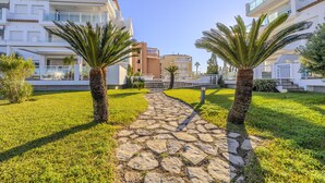 A pathway flanked by palm trees leading to modern buildings with balconies. - LES PORTELLES Nº57 1ª Línea Playa (Denia)
