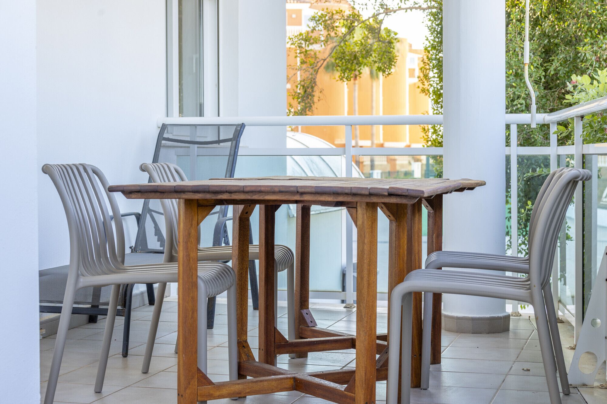 Un balcón con mesa y sillas de madera, ventanas amplias y vista a edificios.