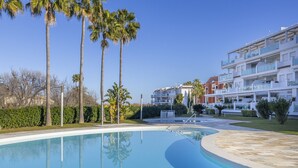 A swimming pool with a curved edge, surrounded by palm trees and a multi-story building with balconies. - LES PORTELLES Nº57 1ª Línea Playa (Denia)