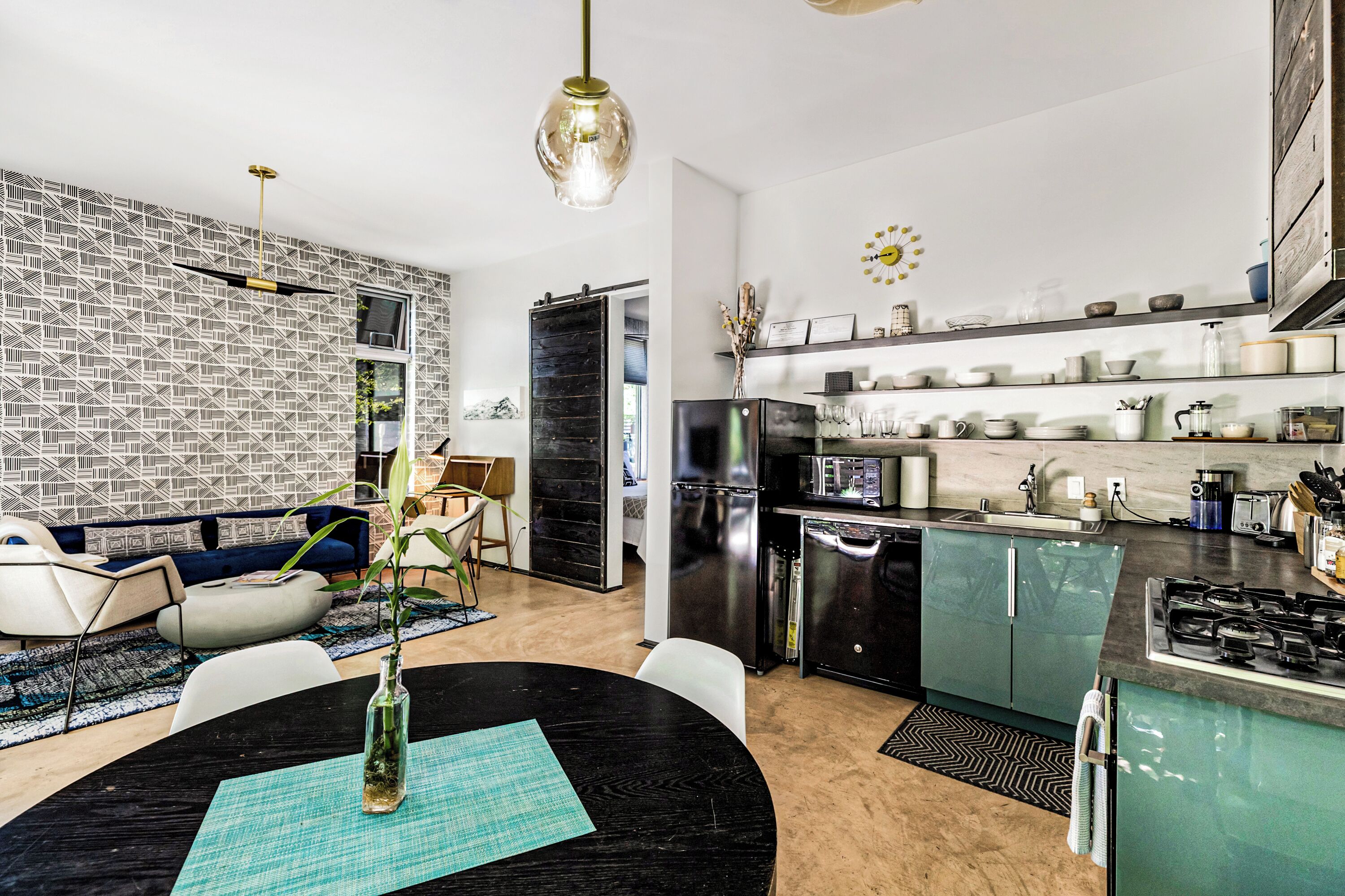 Eine moderne Küche mit einem schwarzen Kühlschrank, Edelstahlgeräten und einer integrierten Kochinsel mit Gasherd. Es gibt einen Essbereich mit einem schwarzen Tisch und Stühlen sowie ein Wohnzimmer mit einem blauen Sofa und einem gemusterten Teppich.