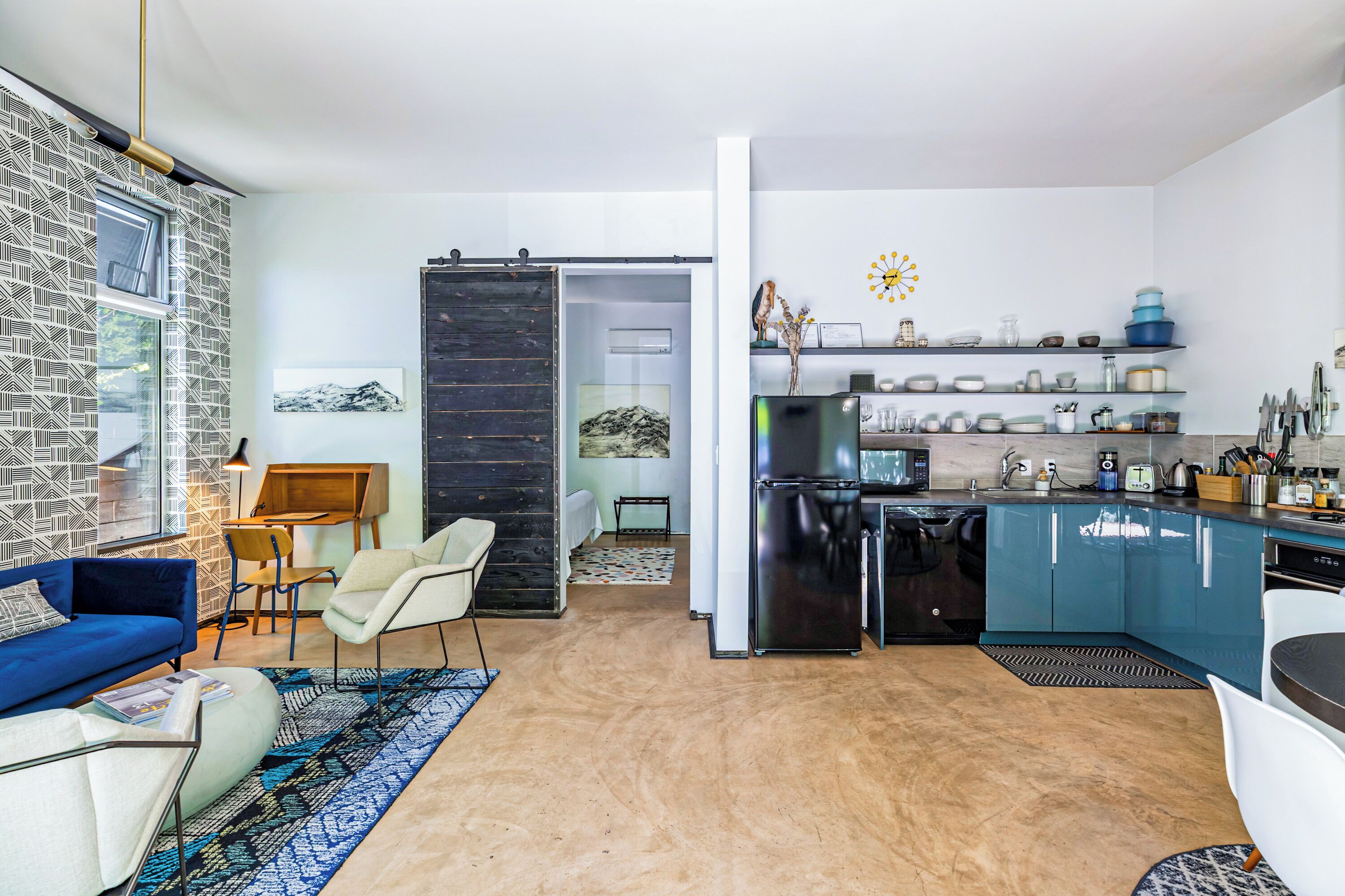 A modern living room with a blue sofa, a white armchair, and a coffee table. There is a kitchen area with blue cabinets and a refrigerator.