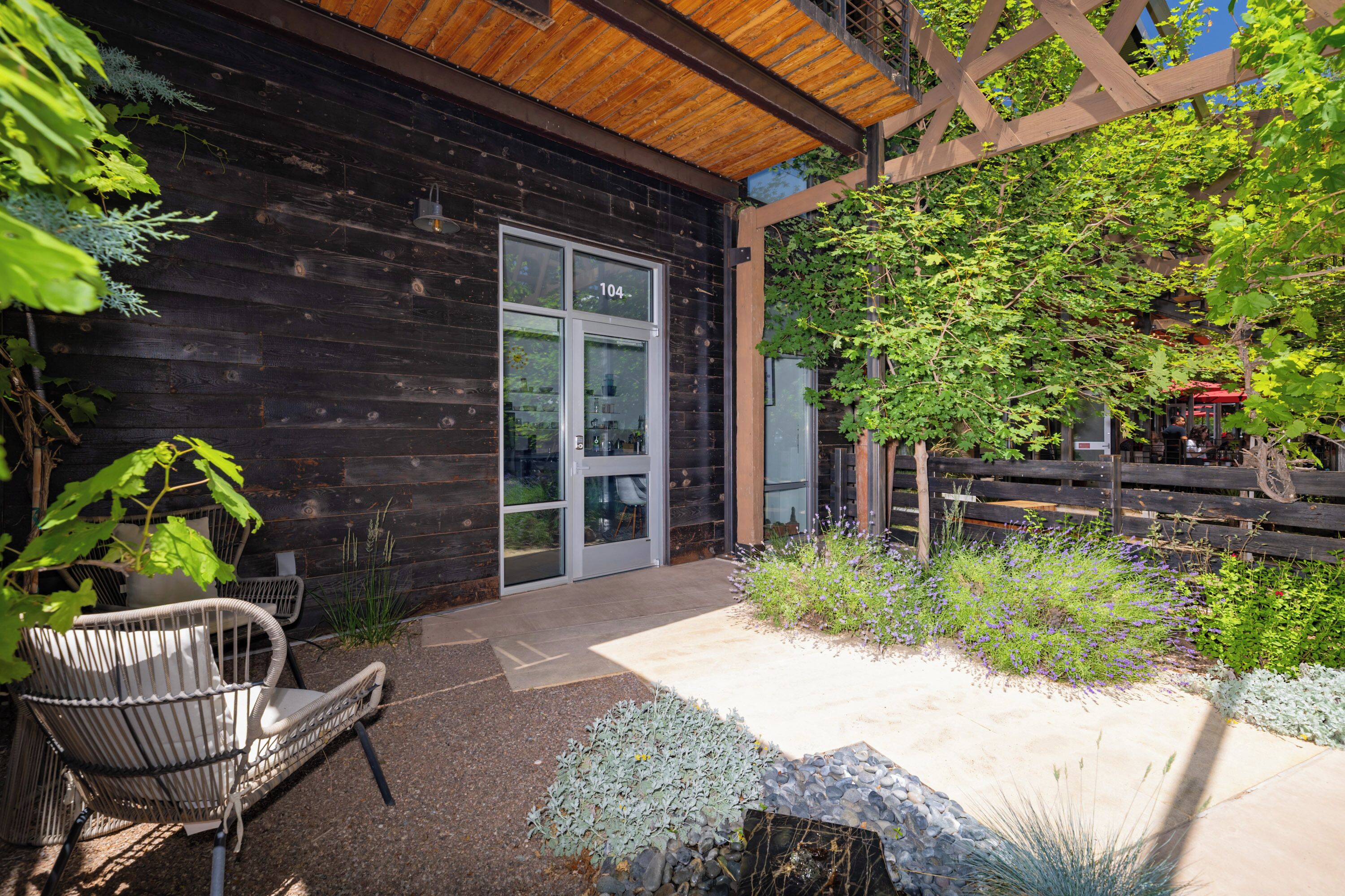 A wooden building with a glass door, a patio with a chair, and a garden with various plants.