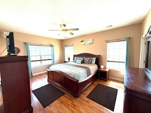 A bedroom with a wooden bed, a ceiling fan, two windows with curtains, a nightstand, and a dresser.