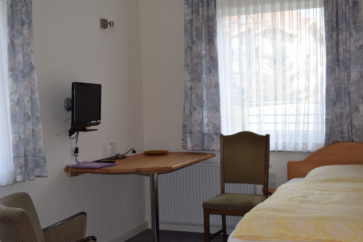 A hotel room with a bed, a desk with a computer, a chair, and a window with curtains.