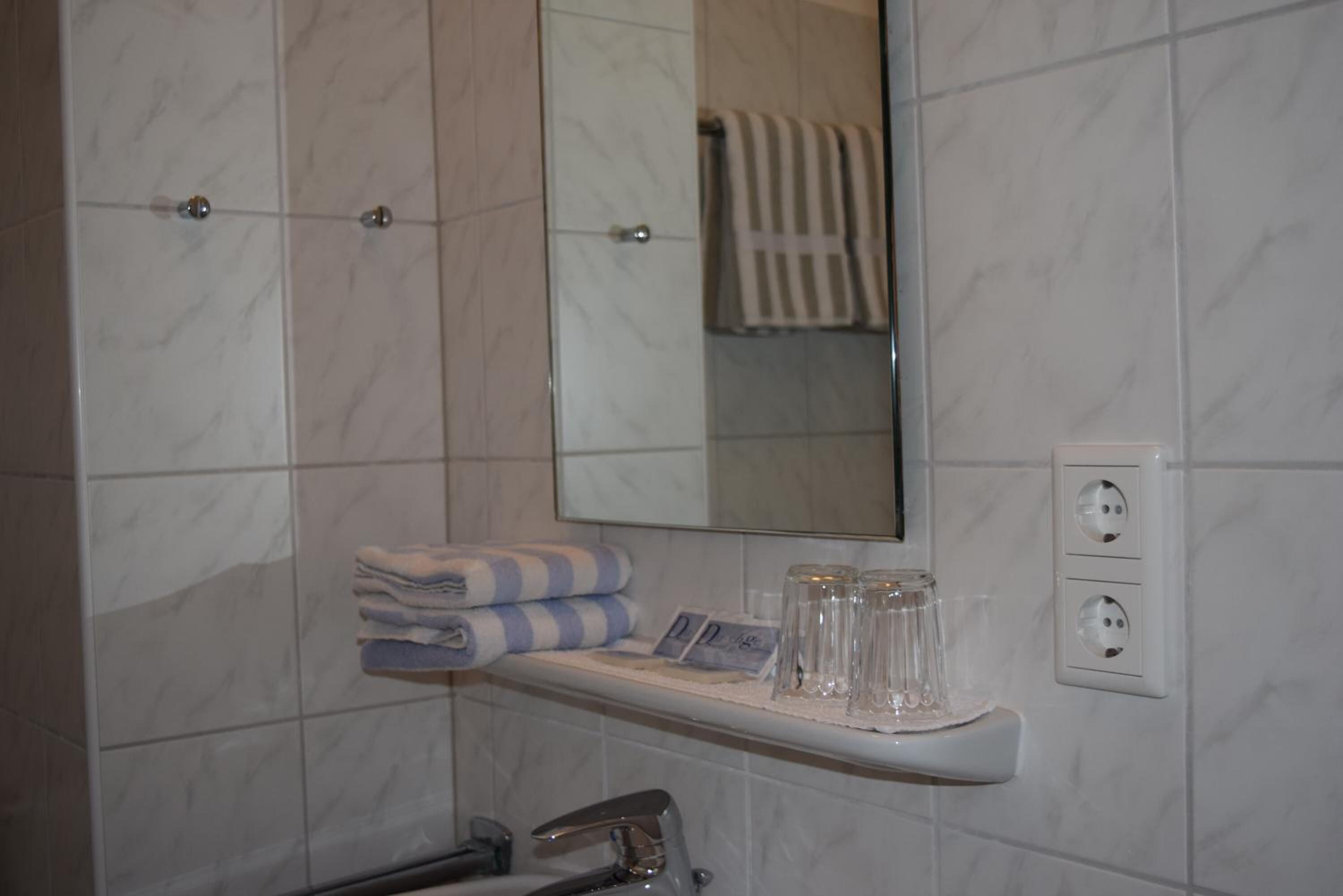A bathroom with a marble wall, a sink with a faucet, a towel rack, a mirror, and a shelf with glasses and a towel.