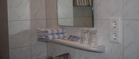 A bathroom with a marble wall, a sink with a faucet, a towel rack, a mirror, and a shelf with glasses and a towel.