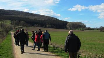 A group of people walking on a dirt path in a rural area with open fields and a forested hill in the background.