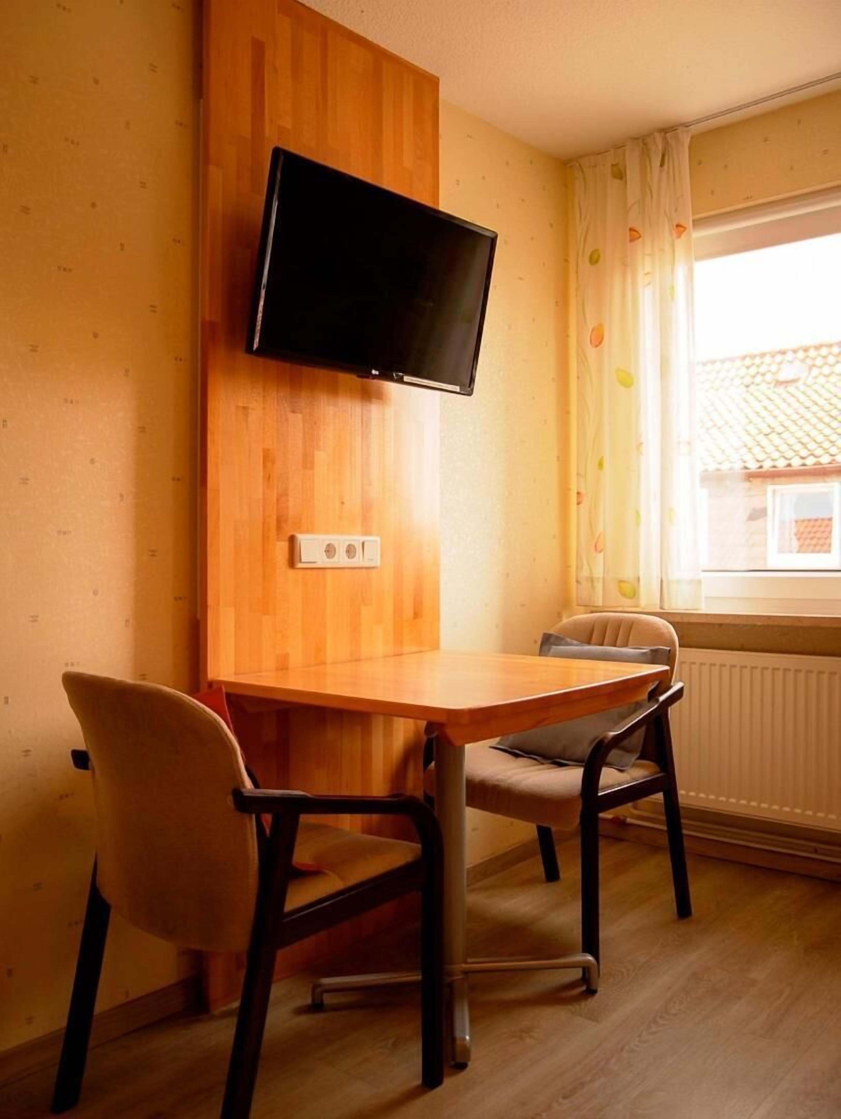 Ein Zimmer mit einem Holzes Esstisch, zwei Stühlen, einem an der Wand befestigten Fernseher und einem Fenster mit Vorhängen.