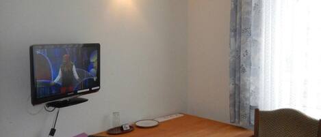 A small room with a single television mounted on the wall, a wooden table, a chair, and a window with curtains.