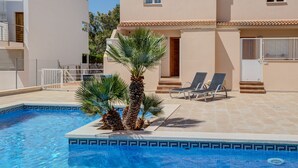 A swimming pool with palm trees, two lounge chairs, and a building with a tiled roof and windows. - Family friendly apartment with pool, close to all amenities and the Pinewalk.  (Pollensa)
