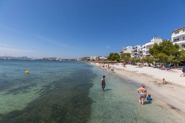 A beach with clear water, people, and buildings in the background.