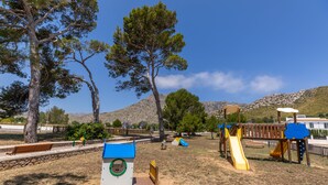 A playground with slides and a bench, surrounded by trees and a mountain in the background. - Family friendly apartment with pool, close to all amenities and the Pinewalk.  (Pollensa)
