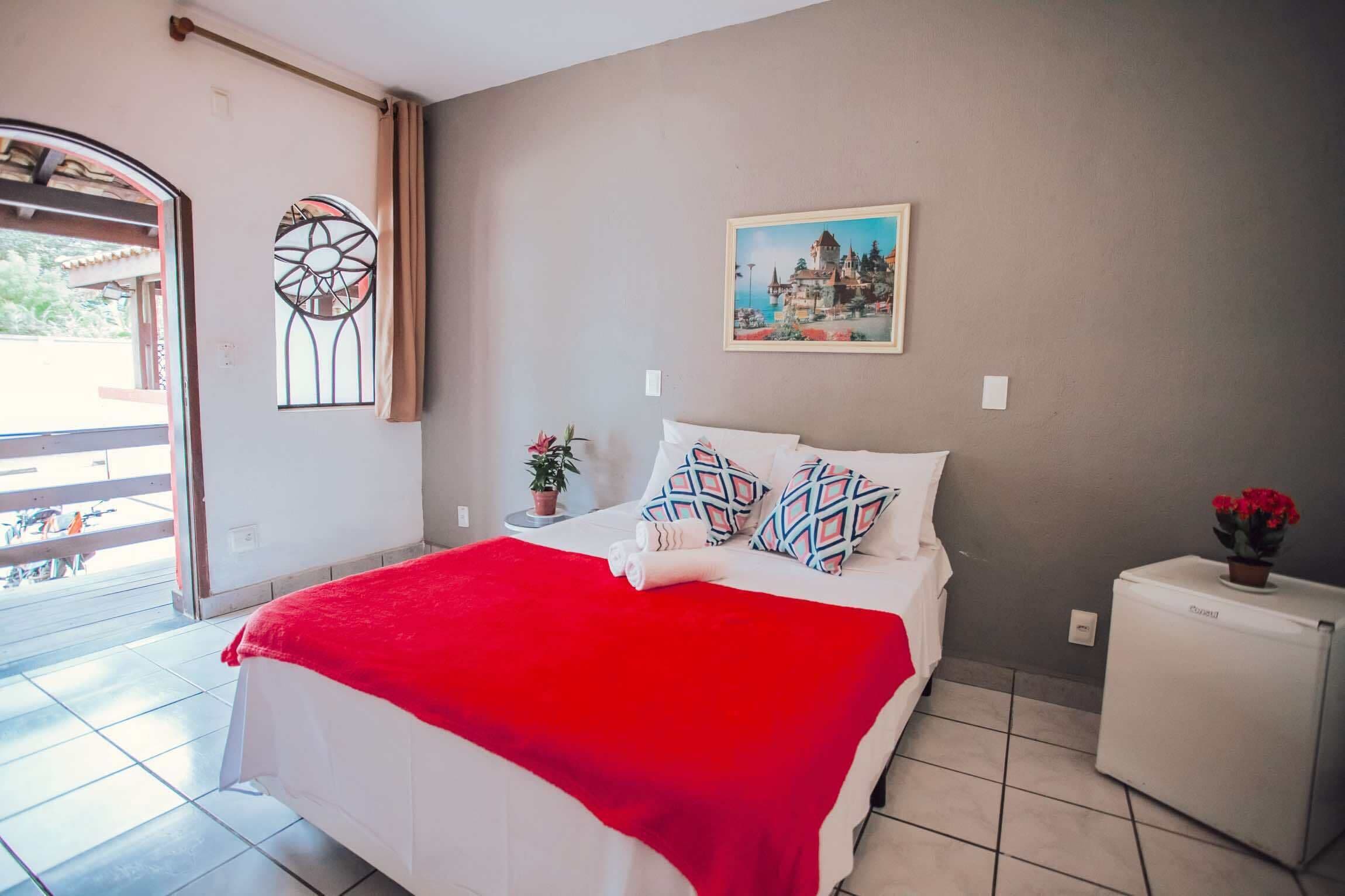 A bedroom with a bed, a red blanket, a small table with a plant, a wall-mounted picture, and a refrigerator.
