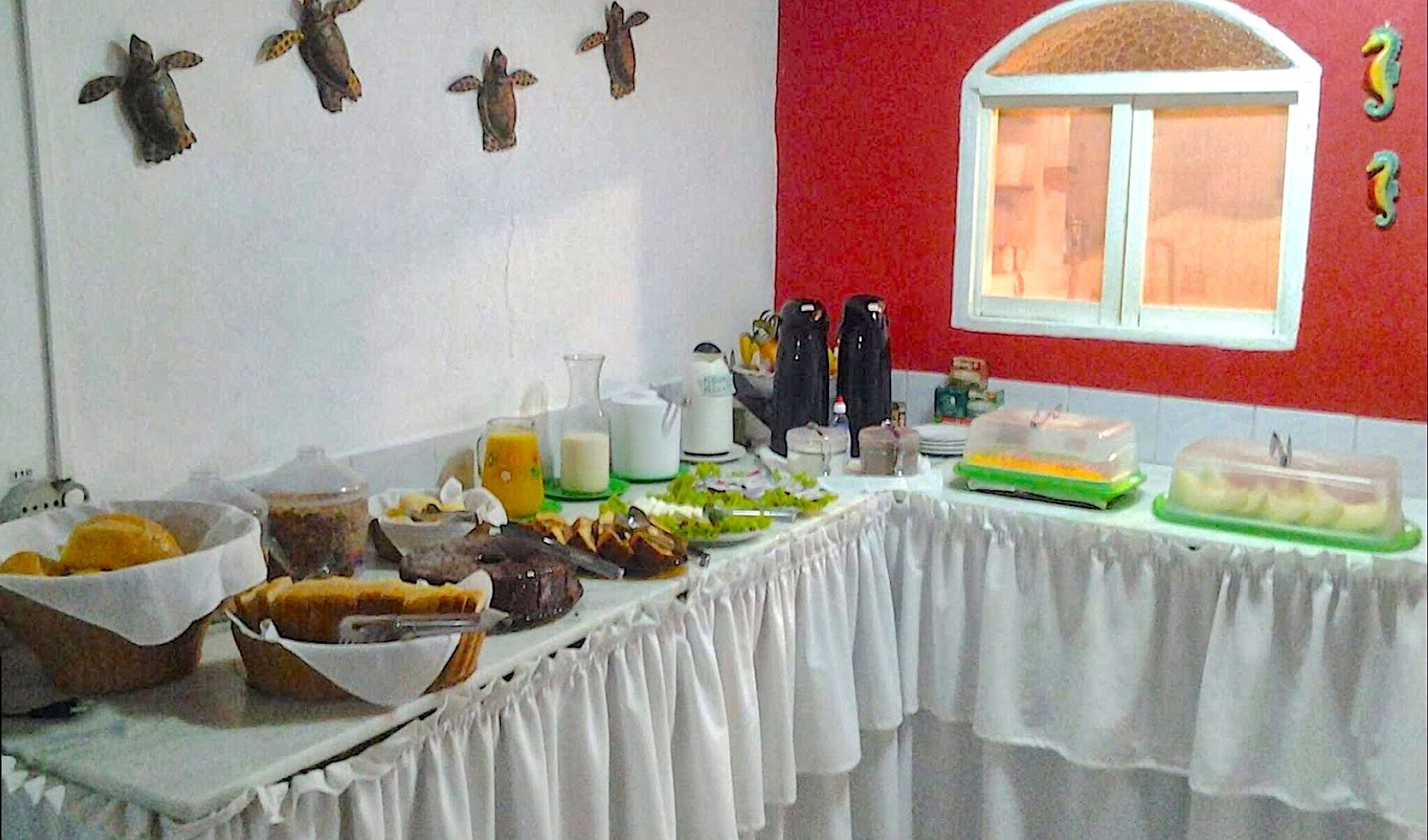 A breakfast buffet with various food items and condiments on a table, set against a red wall with a window.