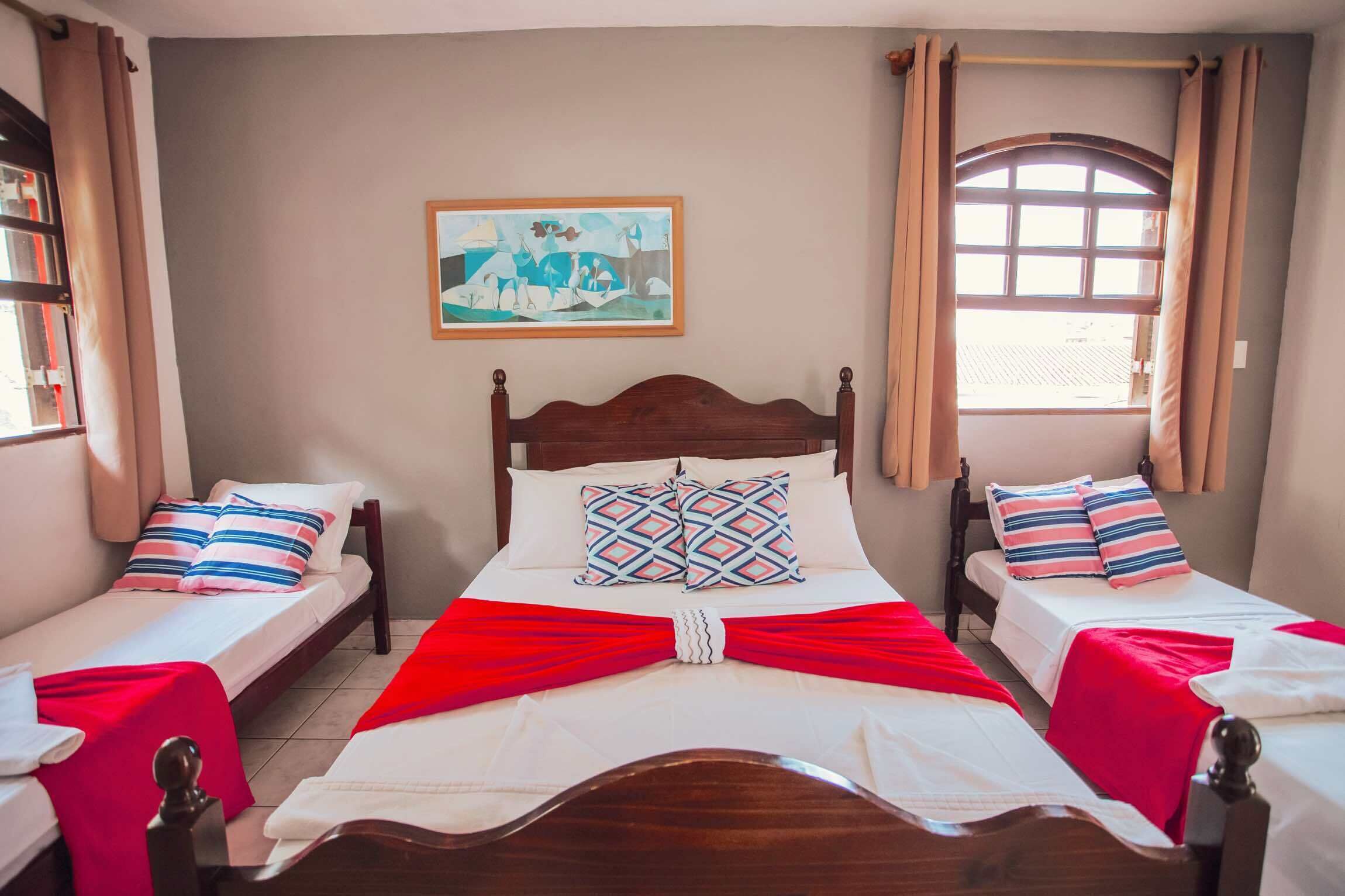Habitación de hotel con una cama grande, dos camas más pequeñas, un cuadro en la pared y una ventana con cortinas.
