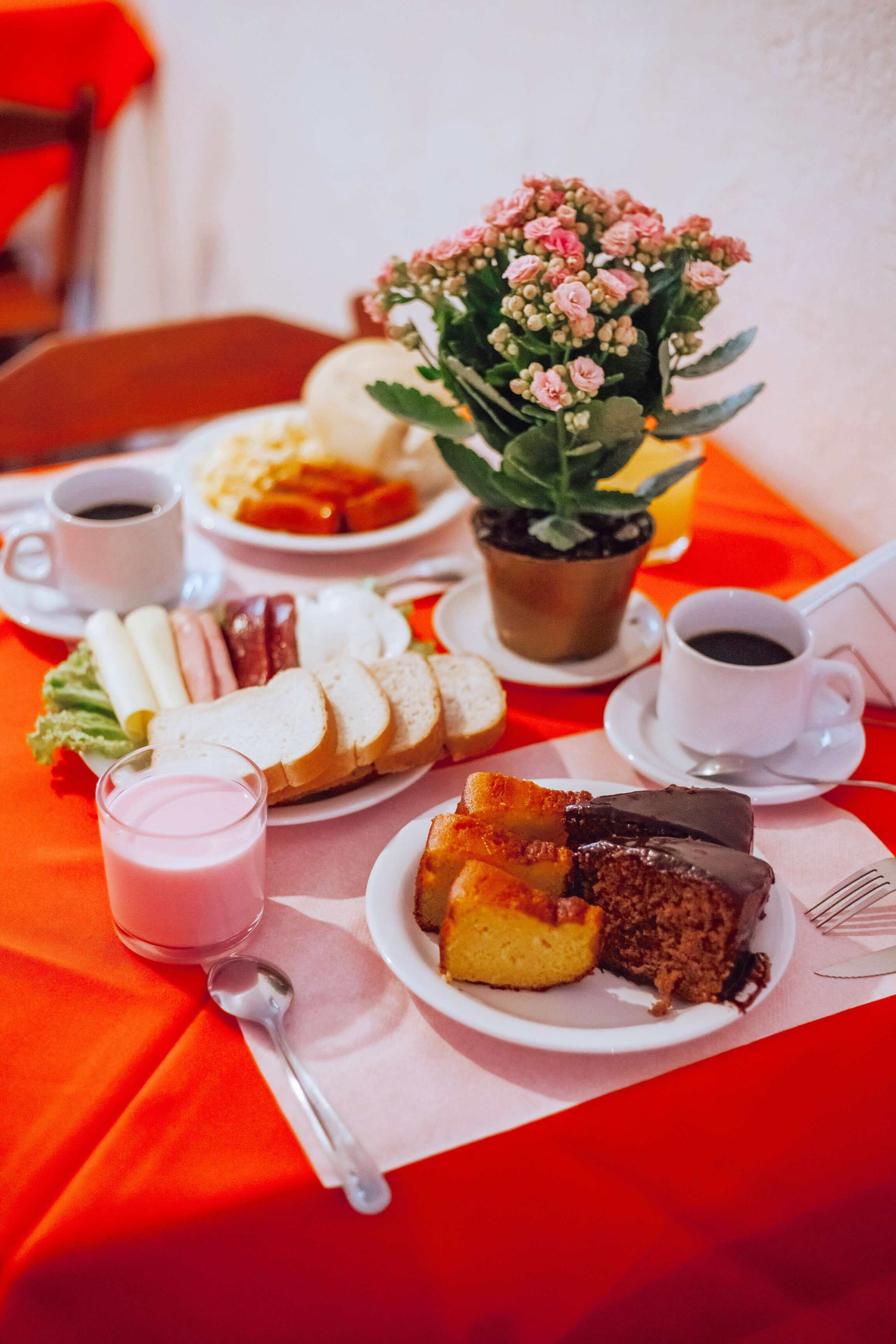 Una mesa con diversos alimentos, como pan, pasteles y un pastel de chocolate, además de una planta en maceta y tazas de café.