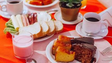 Una mesa con diversos alimentos, como pan, pasteles y un pastel de chocolate, además de una planta en maceta y tazas de café.