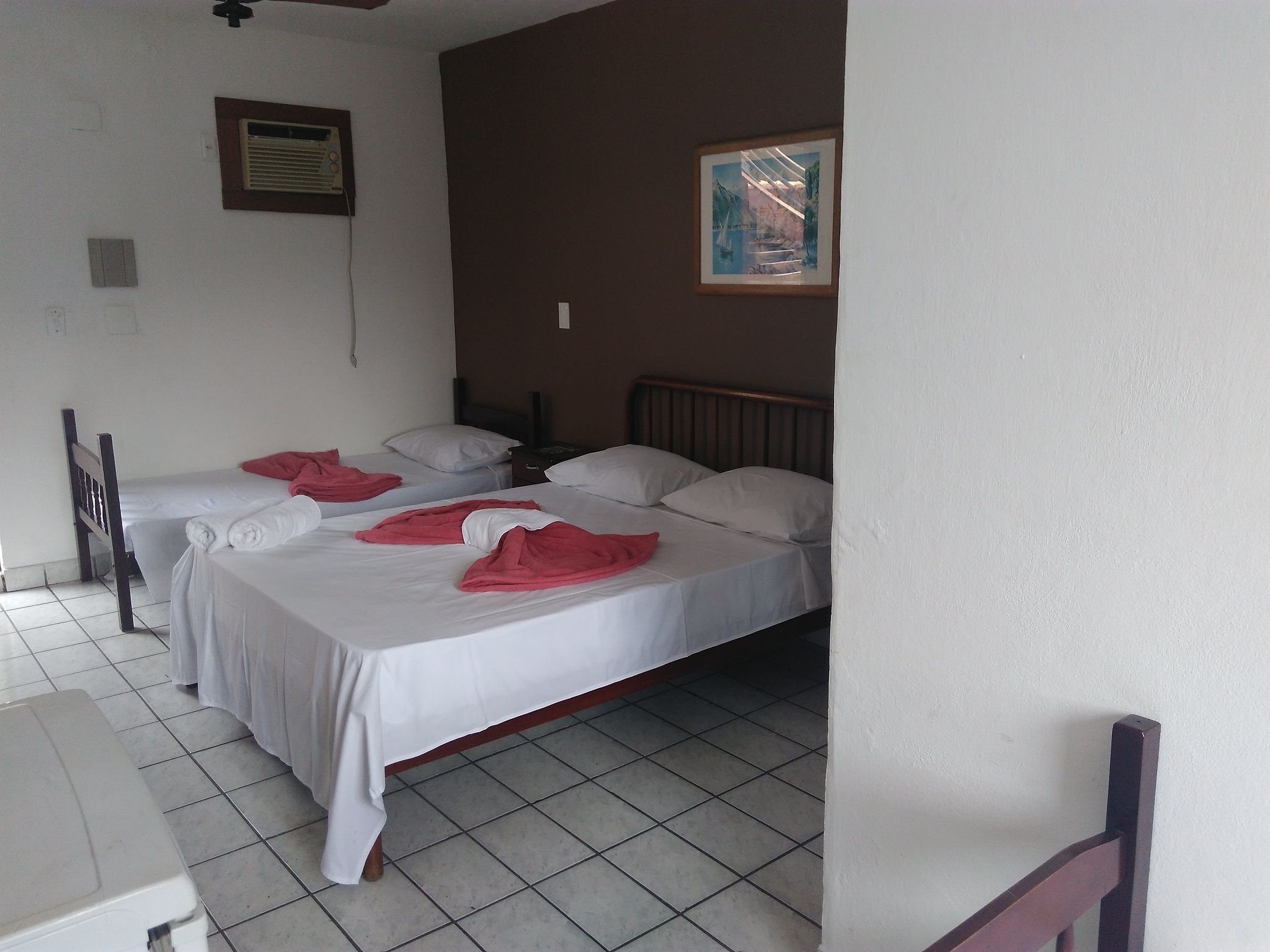 Une chambre d’hôtel avec deux lits, du linge de lit blanc et des serviettes rouges. Il y a un tableau au mur et un ventilateur de plafond.
