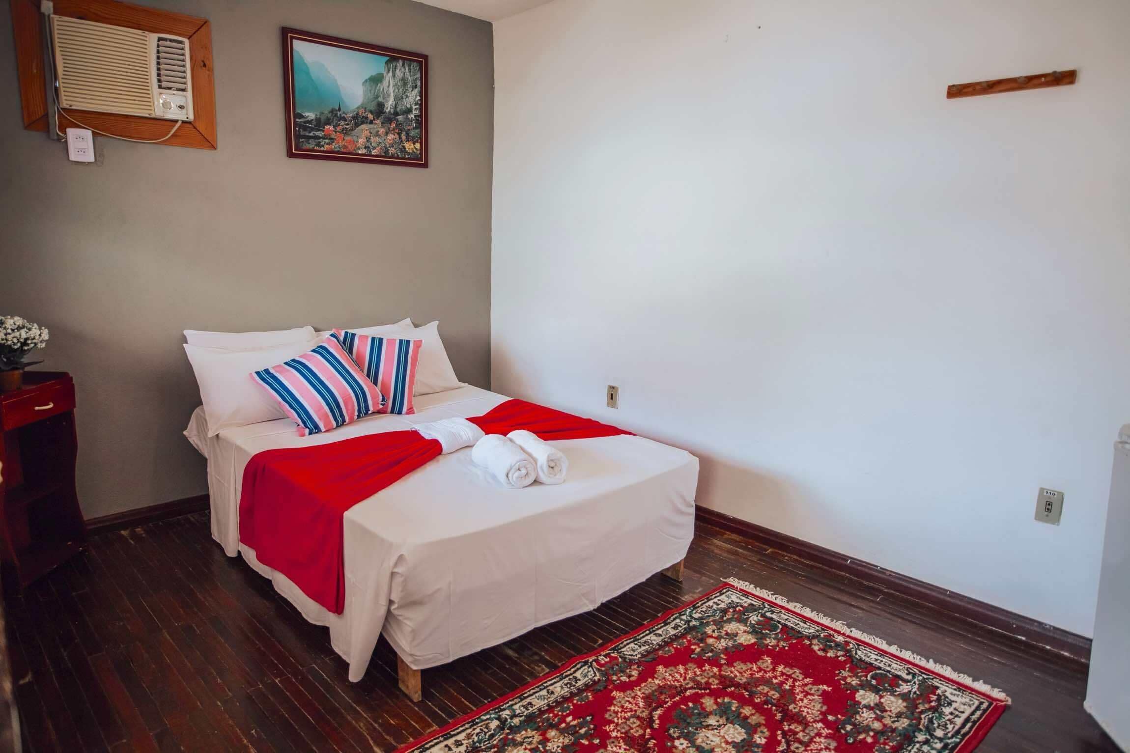 A neatly made bed with a red blanket and striped pillows, a wooden nightstand with a vase of flowers, a red and green patterned rug, a wall-mounted air conditioner, and a framed painting.