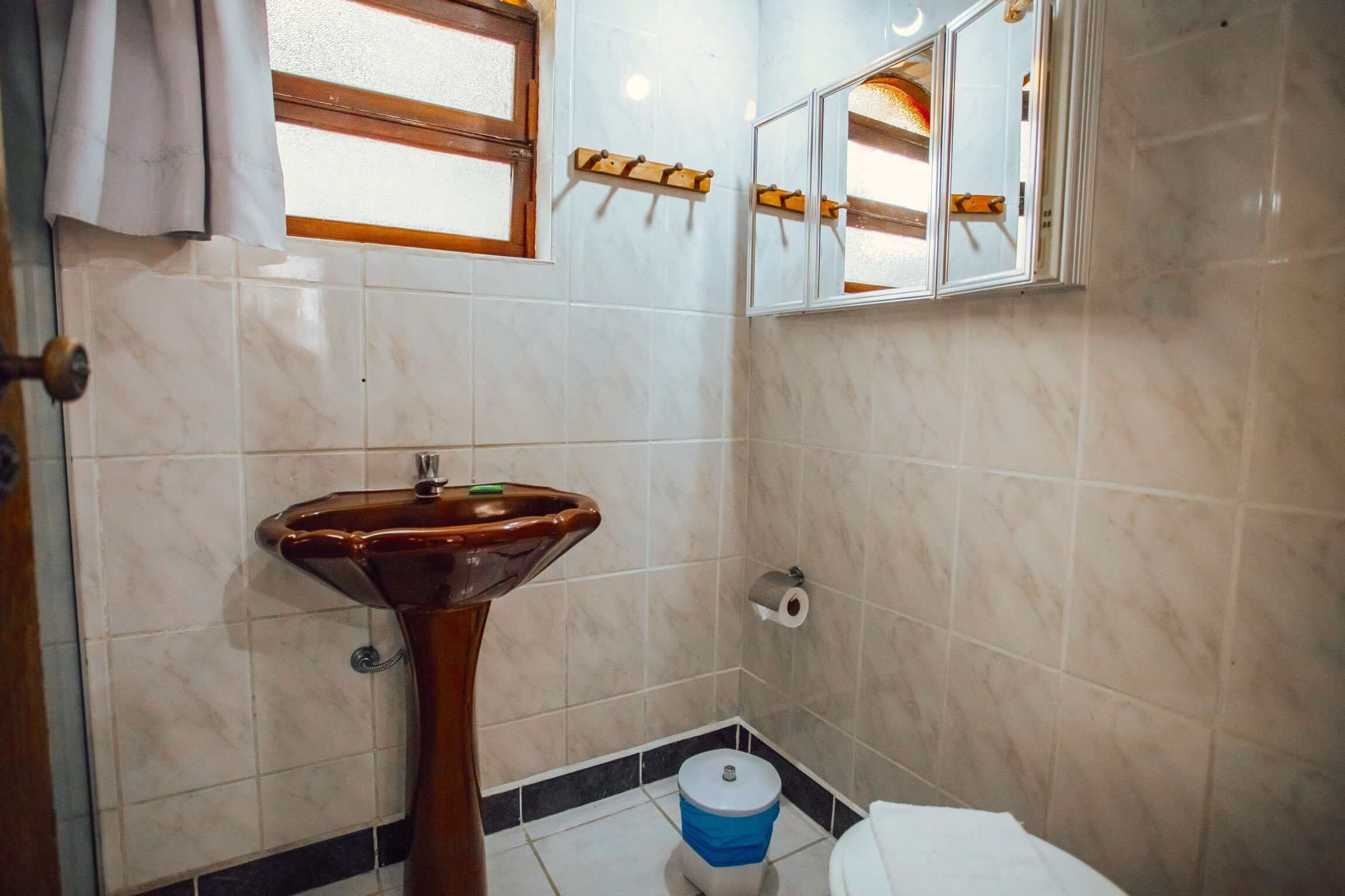 A bathroom with a pedestal sink, a toilet, a blue trash bin, a towel hanging on a door, and a window with a wooden frame.