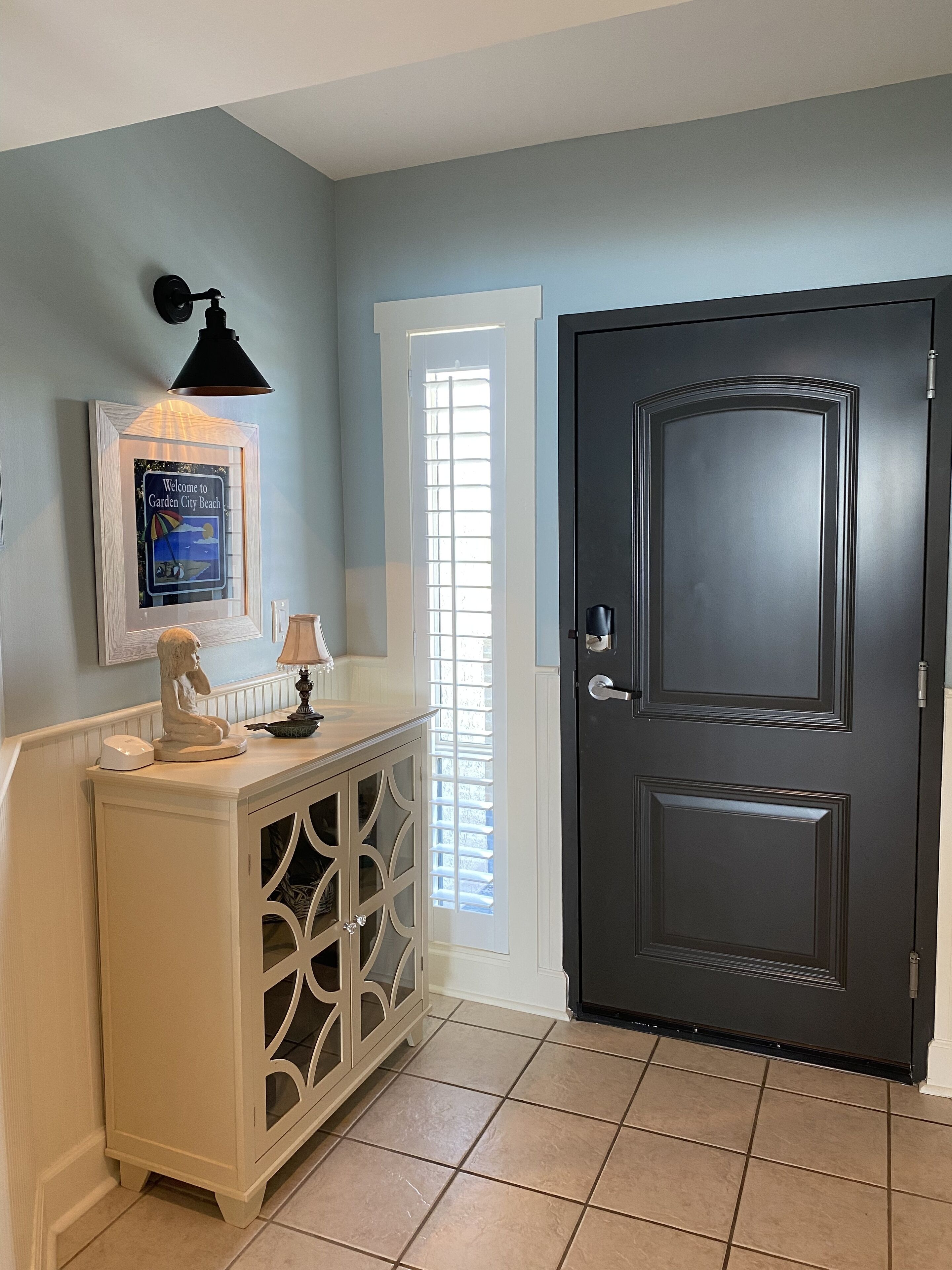 A hallway with a dark door, a white console table with decorative glass doors, a framed welcome sign, and a wall-mounted lamp.