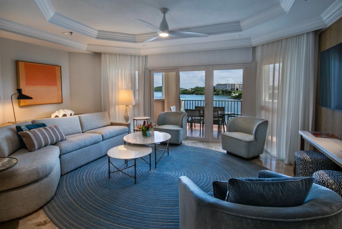 Ein modernes Wohnzimmer mit grauem Sofa, zwei Sesseln, einem Couchtisch und Blick auf das Wasser durch große Fenster.