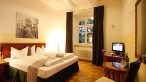 Habitación de hotel con cama, escritorio, silla, televisión, ventana con cortinas y un cuadro en la pared.