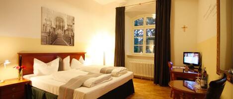 A hotel room with a bed, a desk, a chair, a television, a window with curtains, and a painting on the wall.