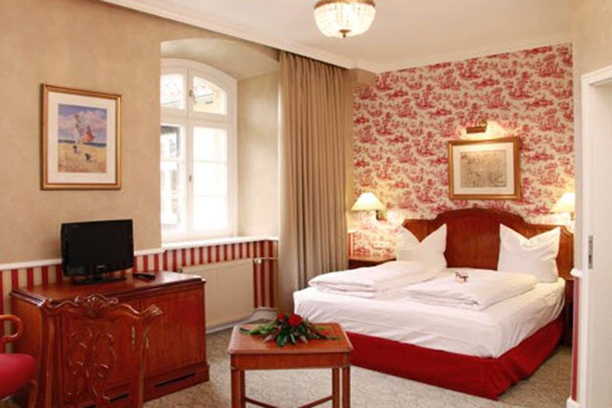 A hotel room with a bed, a desk with a computer, a chair, a window, and a shower area.