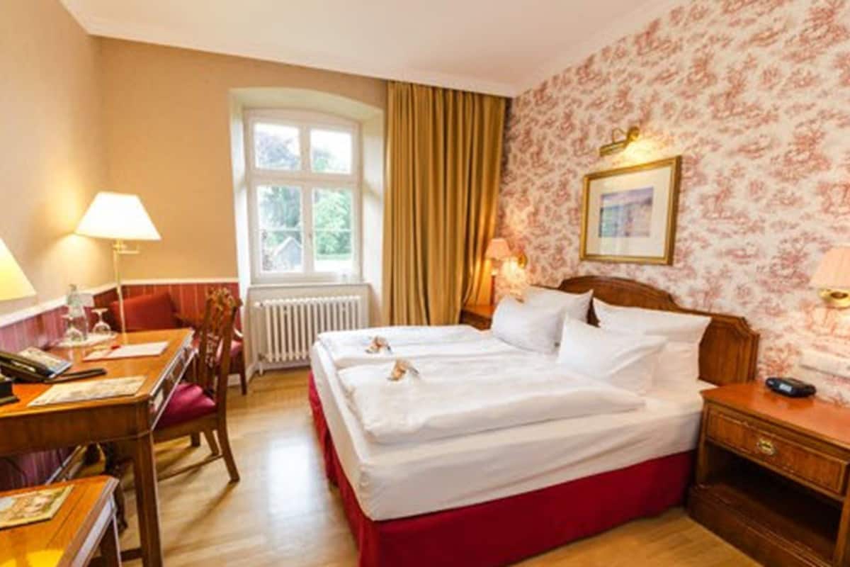 A hotel room with two beds, a desk with a computer, a chair, a window with curtains, and a wall with a floral pattern.