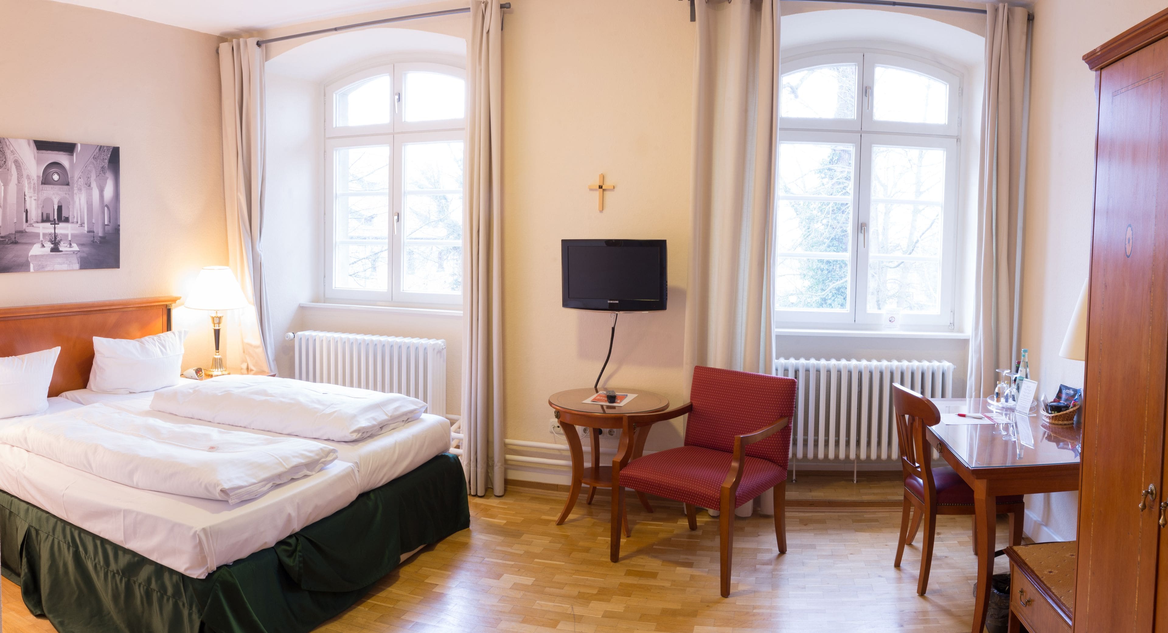 Ett hotellrum med en säng, en tv, en stol, ett skrivbord och ett fönster med gardiner.