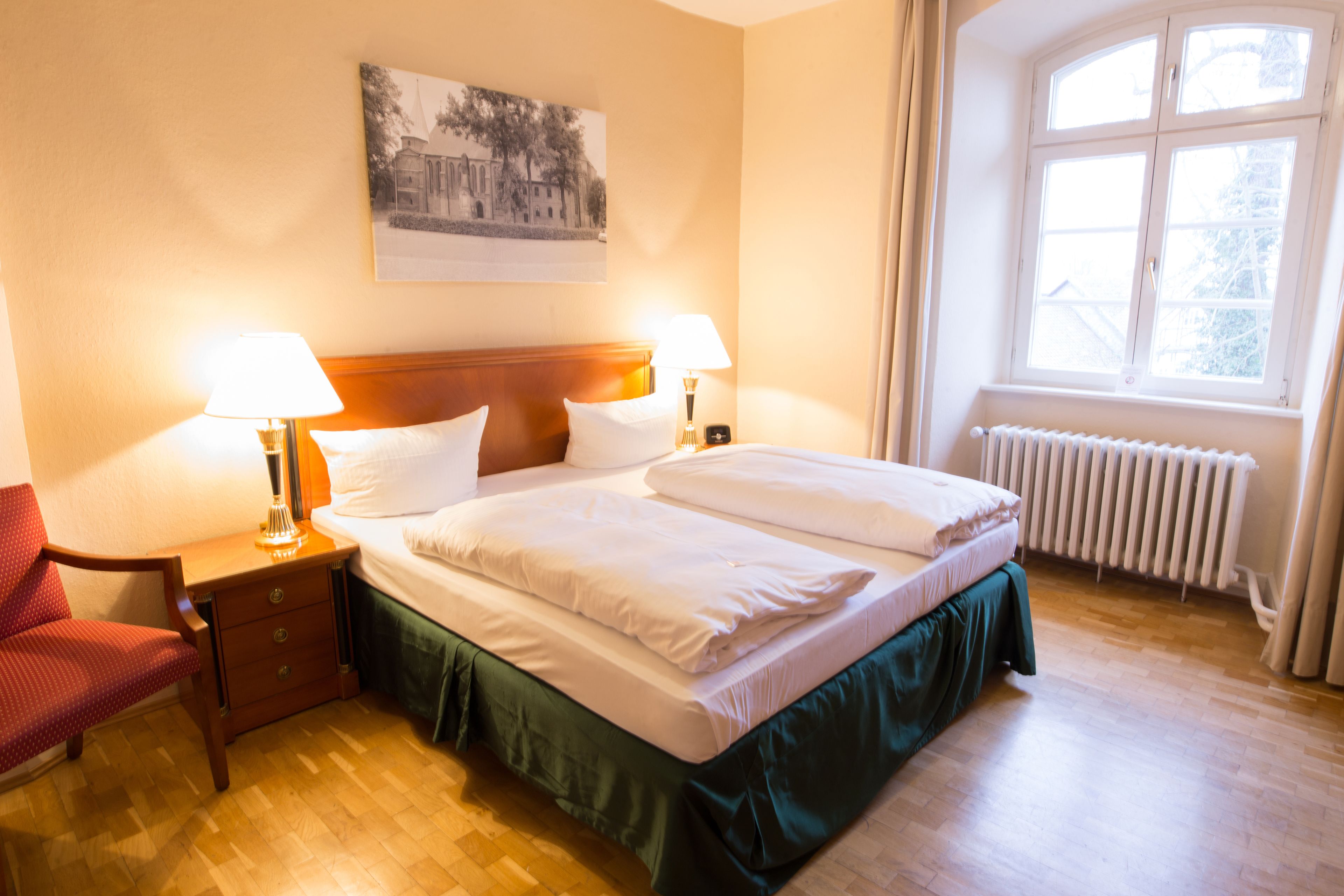 Ein Hotelzimmer mit einem Bett, zwei Nachttischen mit Lampen, einem Sessel, einem Heizkörper und einem Fenster mit Vorhängen.