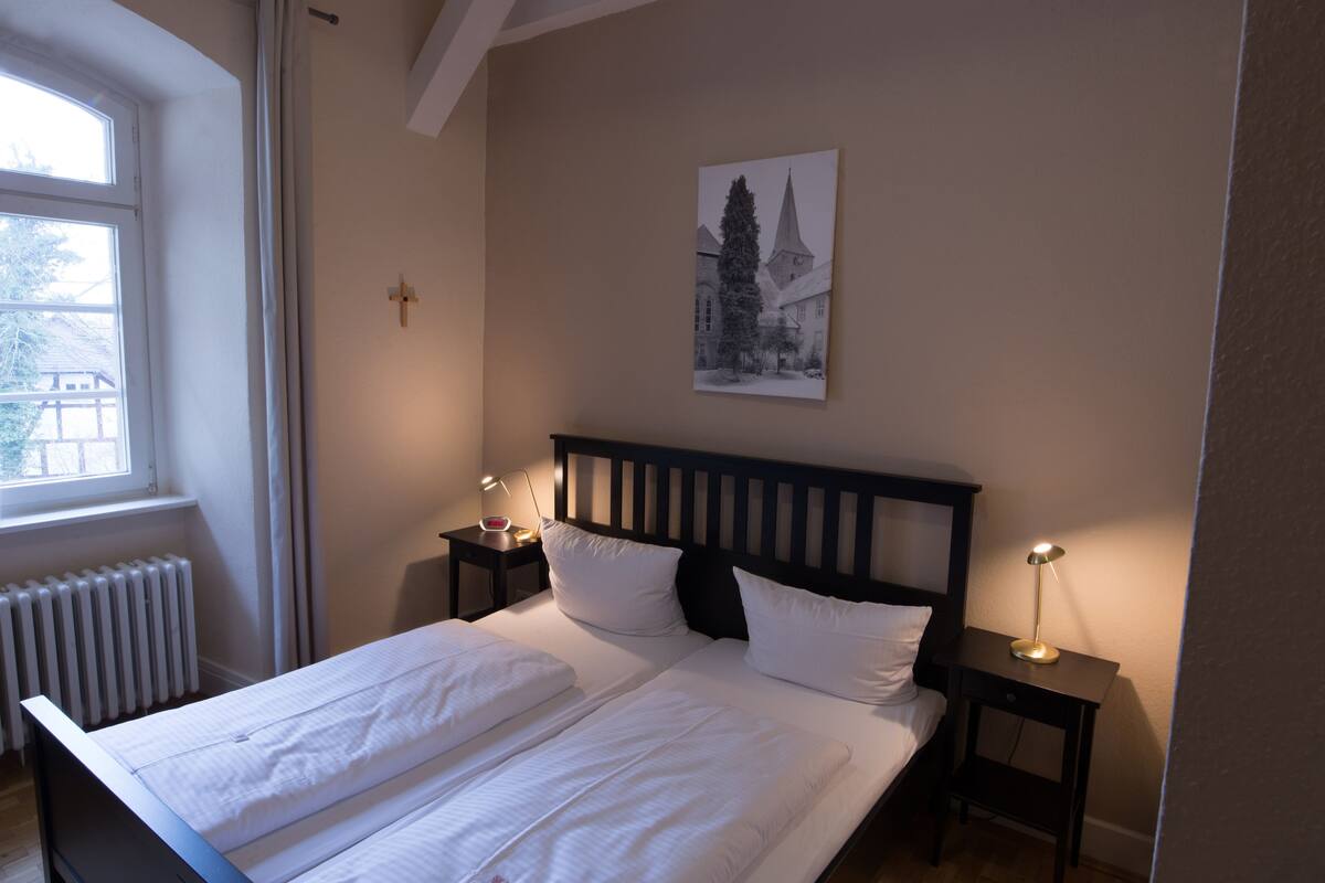 A bedroom with a double bed, two bedside tables with lamps, a radiator, a window, and a wall-mounted picture.