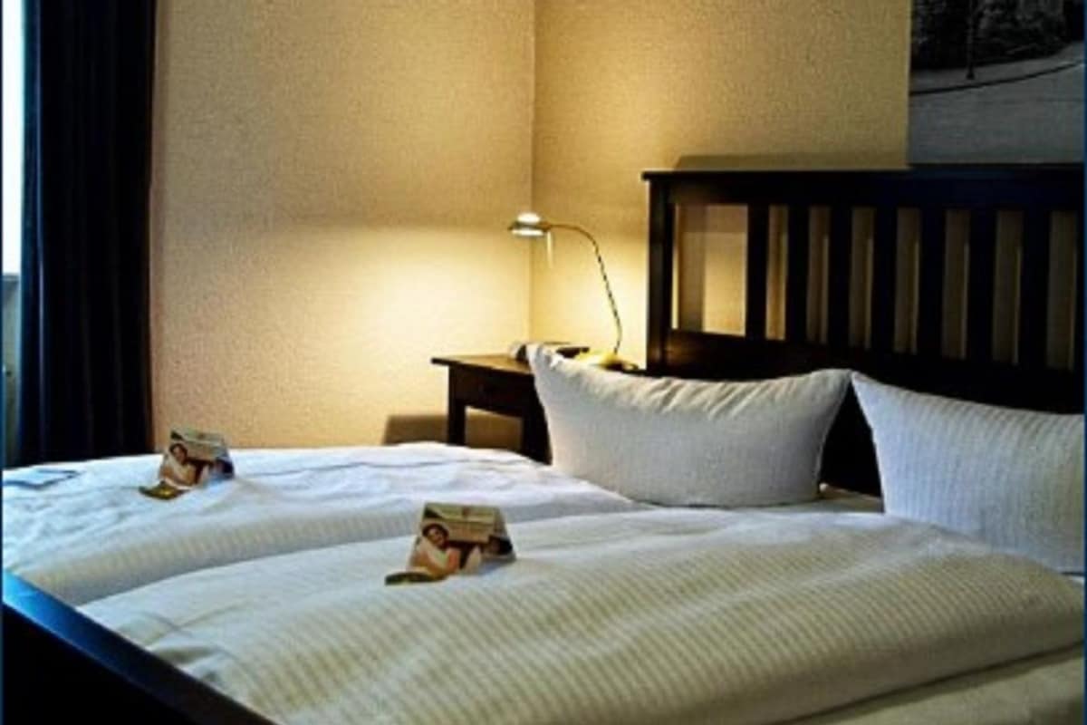 A hotel room with a double bed, a nightstand, a lamp, and a cross on the wall.