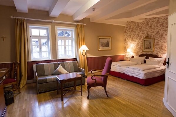 Una habitación de hotel con suelo de madera, una cama con ropa de cama blanca, un sofá, una silla, una mesa y dos ventanas con cortinas.