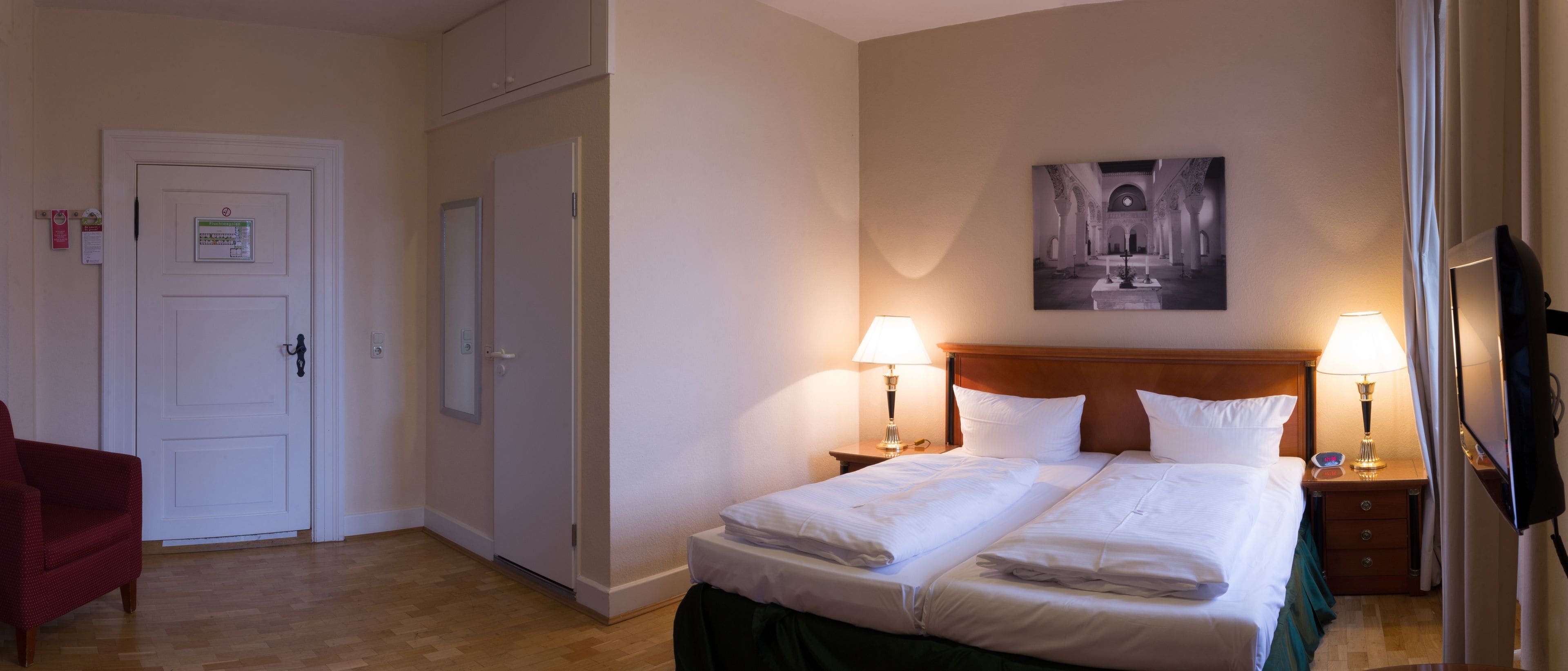 Une chambre d’hôtel avec deux lits, un fauteuil rouge, un tableau au mur et un téléviseur sur le côté droit.