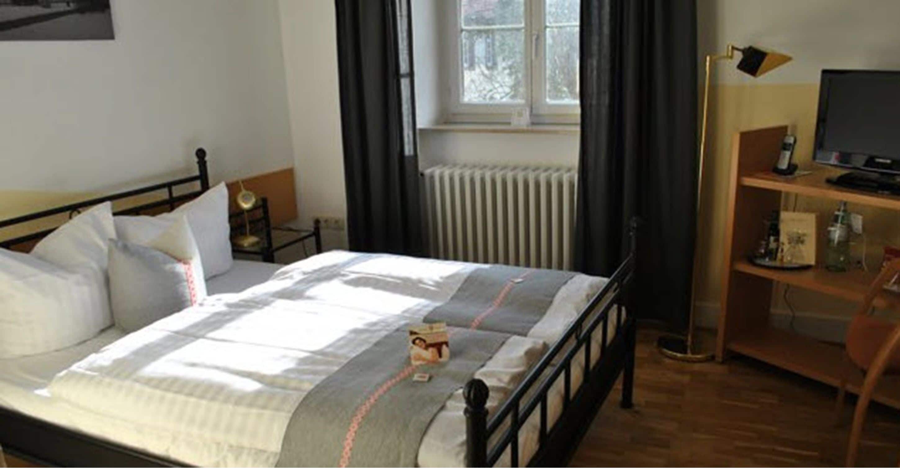 Una cama bien hecha con almohadas y una colcha, una mesita de noche con una lámpara, un radiador y un televisor sobre una estantería de madera.