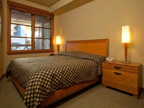 Una cama individual con colcha a cuadros, cabecera de madera, dos lámparas de noche y una mesita con teléfono.