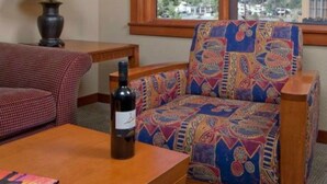 Una habitación con una mesa de madera, un sillón estampado, una lámpara y una botella de vino.
