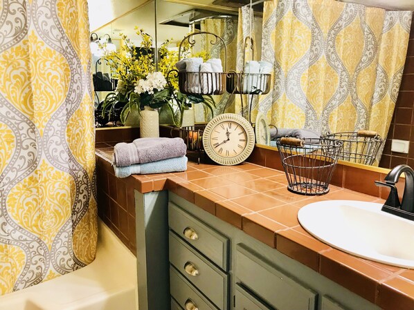 Une salle de bain avec un lavabo, un miroir, une horloge, des serviettes et un panier.