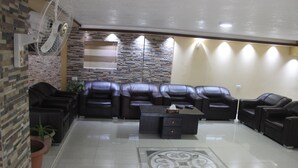 A living room with a sectional sofa, a coffee table, and a fan. There is a stone wall and a window with blinds. - Sara Crown Hotel (Irbid)