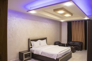 Un dormitorio moderno con cama, mesitas de noche, un área de descanso con sofá y sillón, e iluminación empotrada en el techo.