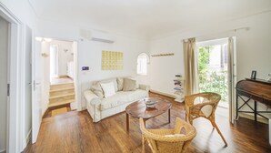 Ein geräumiges Wohnzimmer mit Holzboden, einem weißen Sofa, einem Couchtisch, Korbstühlen, einem Klavier und Blick auf einen Balkon.