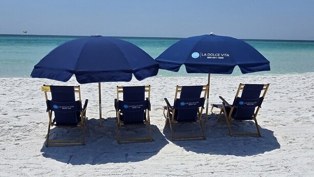Una spiaggia con due ombrelloni blu e quattro sdraio con la scritta 'LA DOLCE VITA' sulla sabbia.