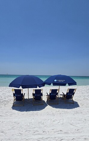 A beach with two blue umbrellas and four lounge chairs labeled "LA DOLCE VITA" on a sandy shore. - Sleeps 8, dog friendly, private beach, beach service for 4! NEW BEACH RAMP! (Miramar Beach)