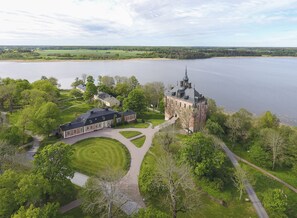 Un edificio grande e histórico con una torre destacada, rodeado de exuberante vegetación y un lago.