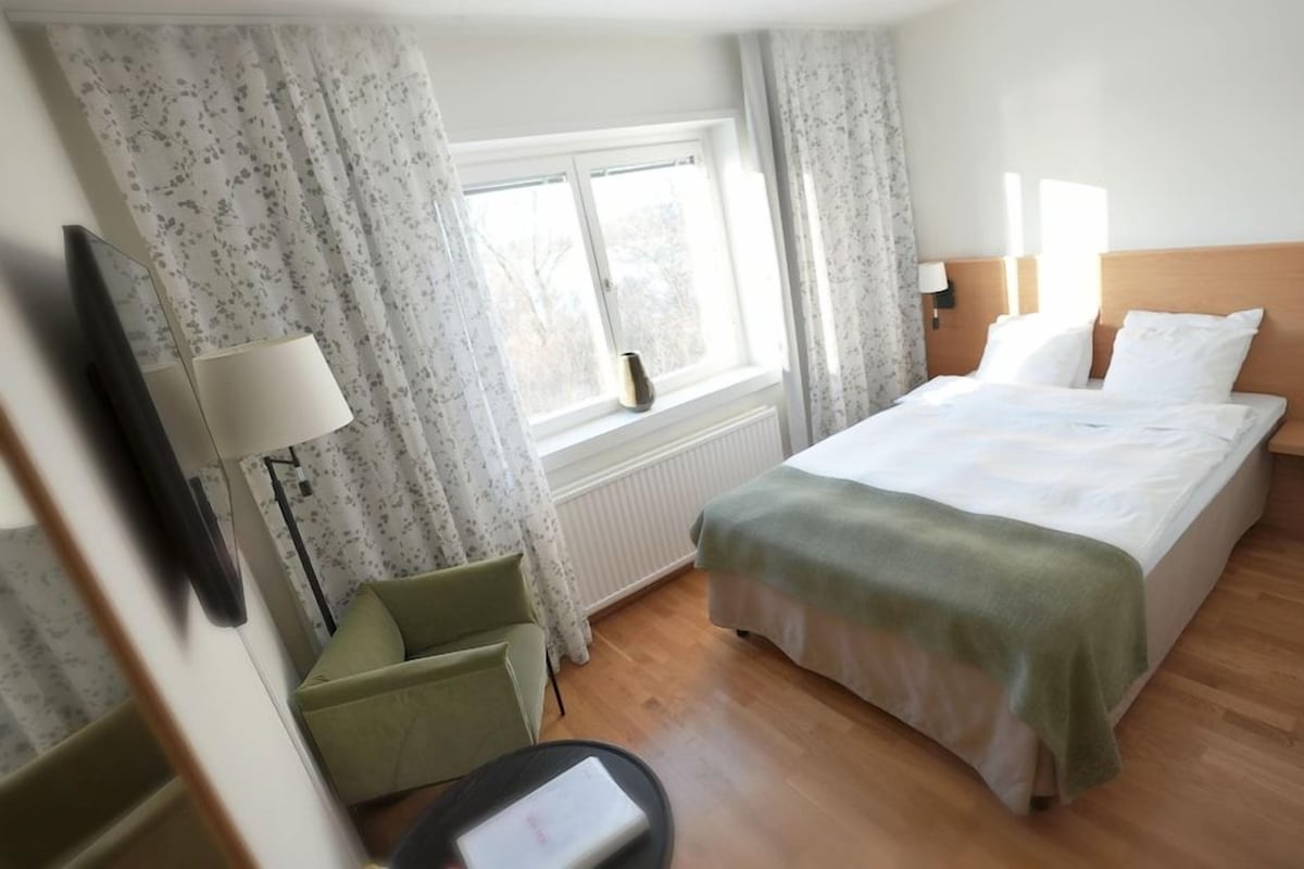 Una habitación de hotel con cama, una silla, una mesa, una lámpara y ventana con cortinas.