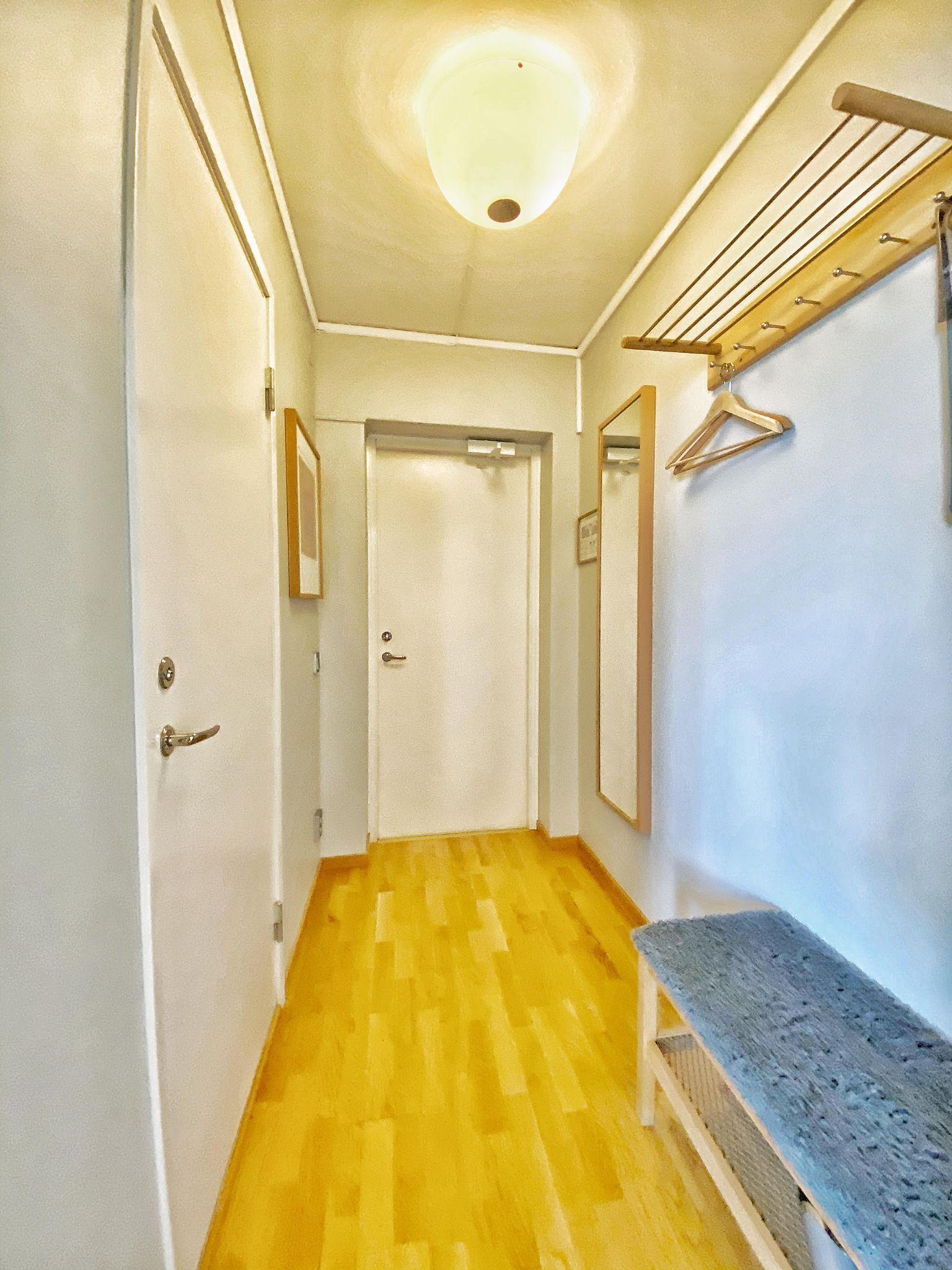 A hallway with a wooden floor, a bench, a drying rack, and a door at the end.