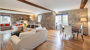 Un vaste espace de vie avec des murs en pierre, des poutres apparentes, un ensemble de canapés, une table à manger avec des chaises et une cheminée en pierre.